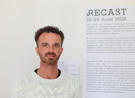 A man with curly hair and a beard smiles at the camera, standing in front of a white exhibition wall.