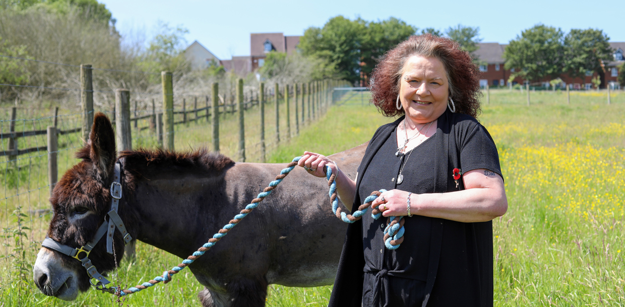 A woman in a black shirt stands smiling in a field, holding a leash attached to a calm donkey.