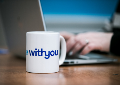 A white mug with the phrase "WithYou" in focus on a desk, with blurry hands typing on a laptop in the background.