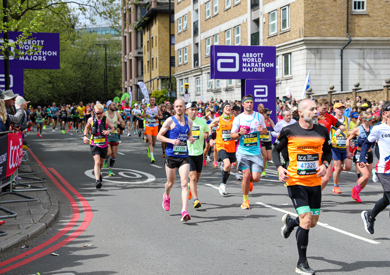 Runner wearing a WithYou vest participating in a city marathon, with spectators along the barricaded course. signs and banners are visible.