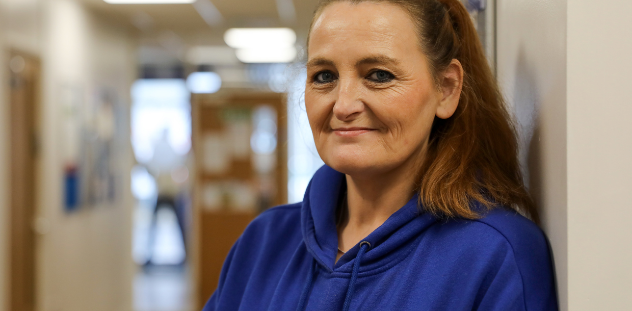 A middle-aged woman, wearing a blue hoodie, stands near a hallway wall, smiling gently towards the camera.