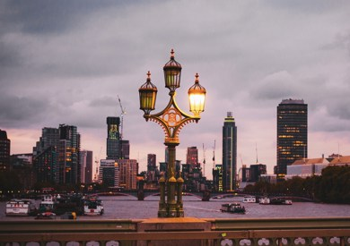 An ornate street lamp in lambeth illuminated at dusk on a bridge, with a backdrop of a city skyline under a cloudy pink and blue sky.