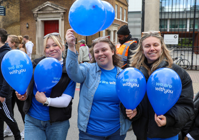 Three women in blue shirts hold blue balloons with "we are with you" printed on them, smiling joyfully during an outdoor event.