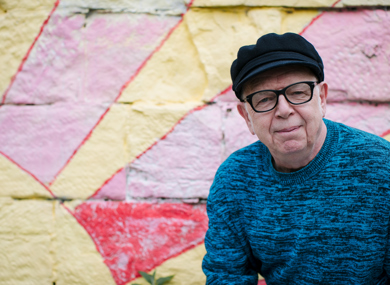 An smiling elderly man in glasses and a black cap. He sits before a colourfully painted wall.