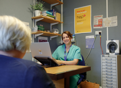 A female nurse in scrubs sits at a desk with a laptop, smiling at an elderly patient whose back is to the camera, in a clinic office decorated with informational posters.