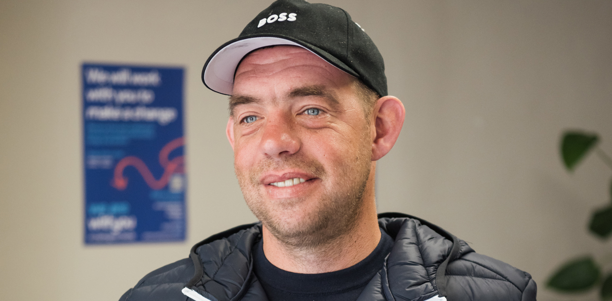 A smiling man wearing a black jacket and cap stands indoors, with a blue WithYou notice board visible in the background.