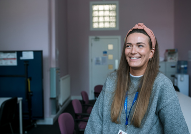 A smiling woman wearing a gray sweater and pink headband sits at a desk in a modern office with chairs and computers in the background.