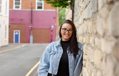 A cheerful young woman with glasses, wearing a denim jacket, stands by a stone wall on a street, with a red building in the background.