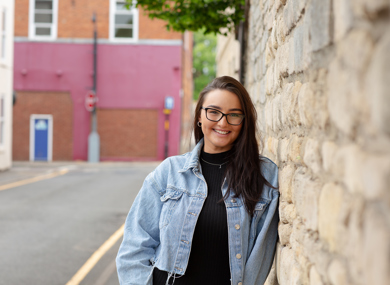 A cheerful young woman with glasses, wearing a denim jacket, stands by a stone wall on a street, with a red building in the background.