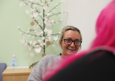 A woman with blonde hair and glasses smiling in a conversation with another person with pink hair. A decorative tree is in the background.