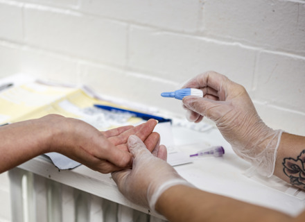 Healthcare worker in gloves performing a blood test on a patient's finger in a clinical setting, with medical supplies around.