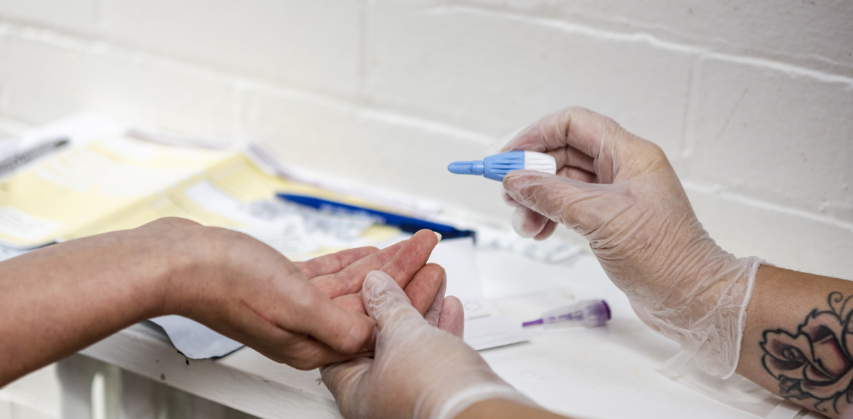 Healthcare worker in gloves performing a blood test on a patient's finger in a clinical setting, with medical supplies around.