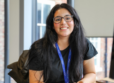 A smiling woman with dark hair and glasses, wearing a black shirt and a blue lanyard, sitting in an office with windows in the background.