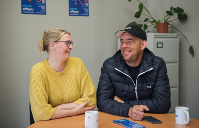A man and a woman smiling at each other while sitting at a table with coffee mugs, pamphlets, and a plant in the background inside an office.