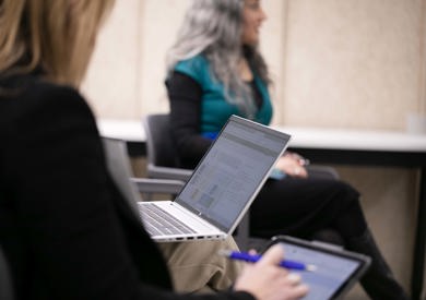 A person sits at a conference table in focus, working on a laptop. Another person in the background is blurred.