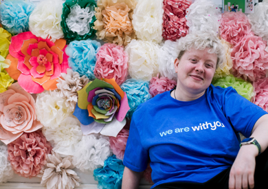 A woman in a blue t-shirt sits smiling in front of a colorful background of assorted paper flowers in various sizes and colors.