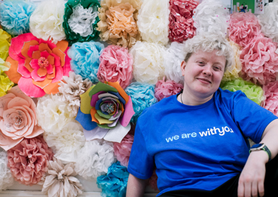 A woman in a blue t-shirt sits smiling in front of a colorful background of assorted paper flowers in various sizes and colors.