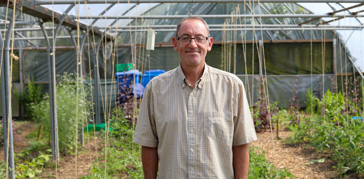 A middle-aged man smiling at the camera, standing in a greenhouse with various plants under a structure with transparent roofing.