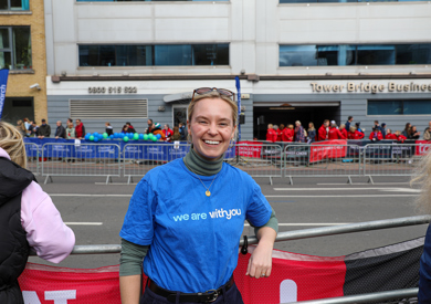 A joyful woman in a blue "we are with you" t-shirt stands behind a red barrier at an outdoor event, with bystanders and blue banners in the background.