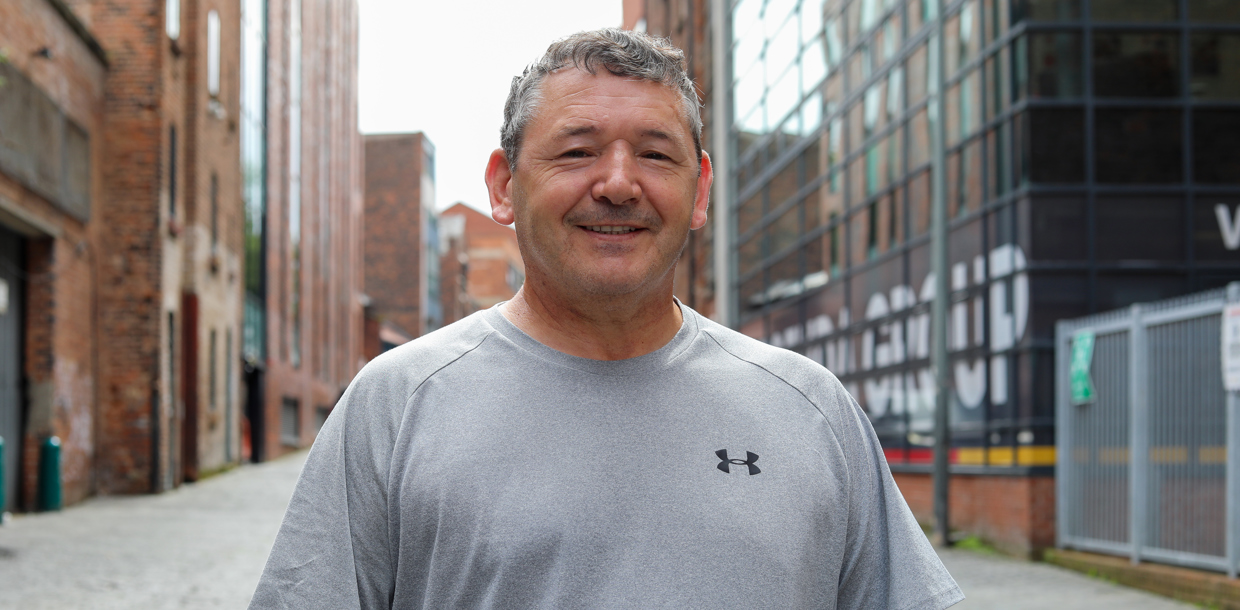 A smiling middle-aged man wearing a grey t-shirt stands in an urban alley with brick buildings and modern structures in the background.