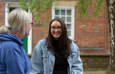 A young woman in glasses and a denim jacket smiles while engaging in a conversation with older woman, seen from the back, in front of a brick building.