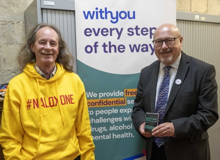 WithYou worker Kevin stood next to Grahame Morris MP. Both are smiling. Grahame Morris is holding a naloxone kit. In the background is a WithYou banner