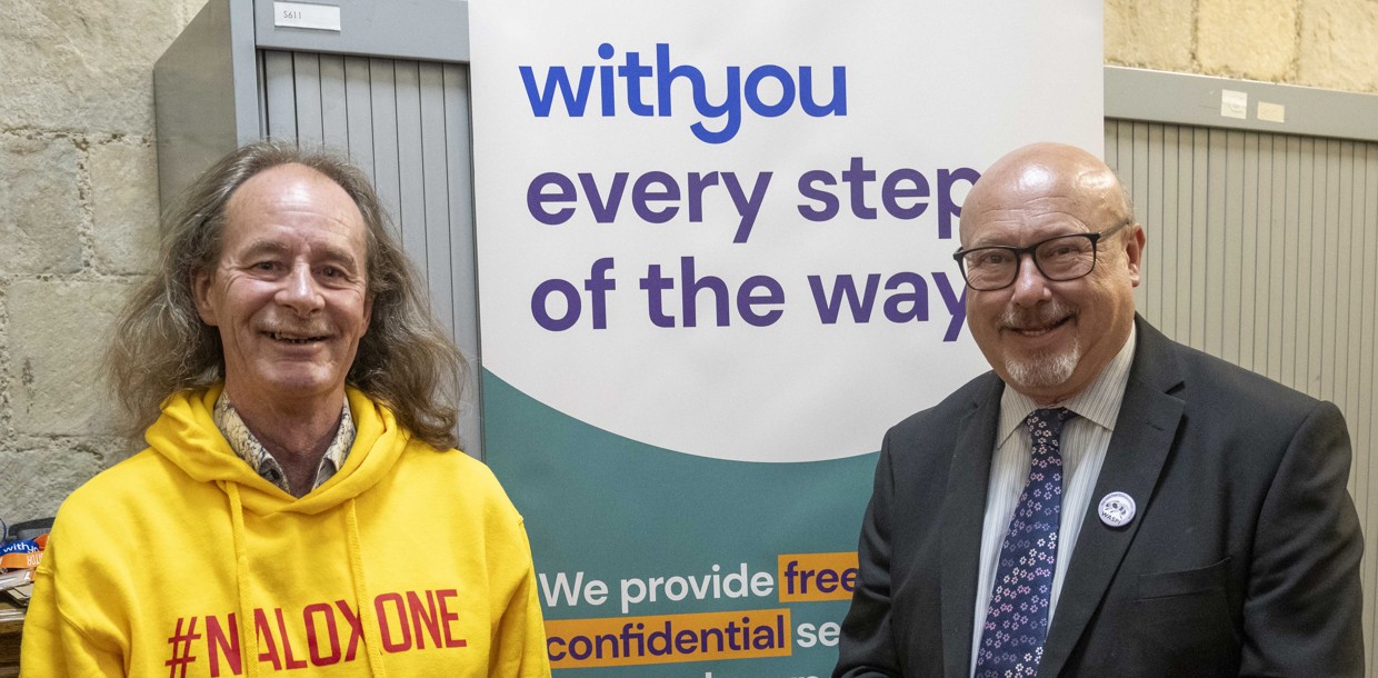 WithYou worker Kevin stood next to Grahame Morris MP. Both are smiling. Grahame Morris is holding a naloxone kit. In the background is a WithYou banner