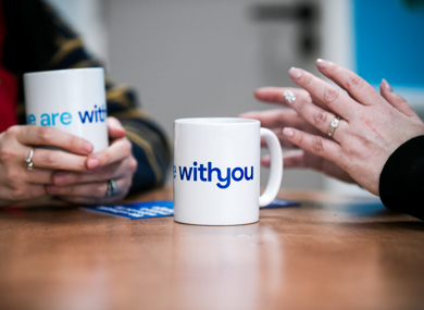 Two people sitting at a table, holding mugs with the text "WithYou" visible on one of them.